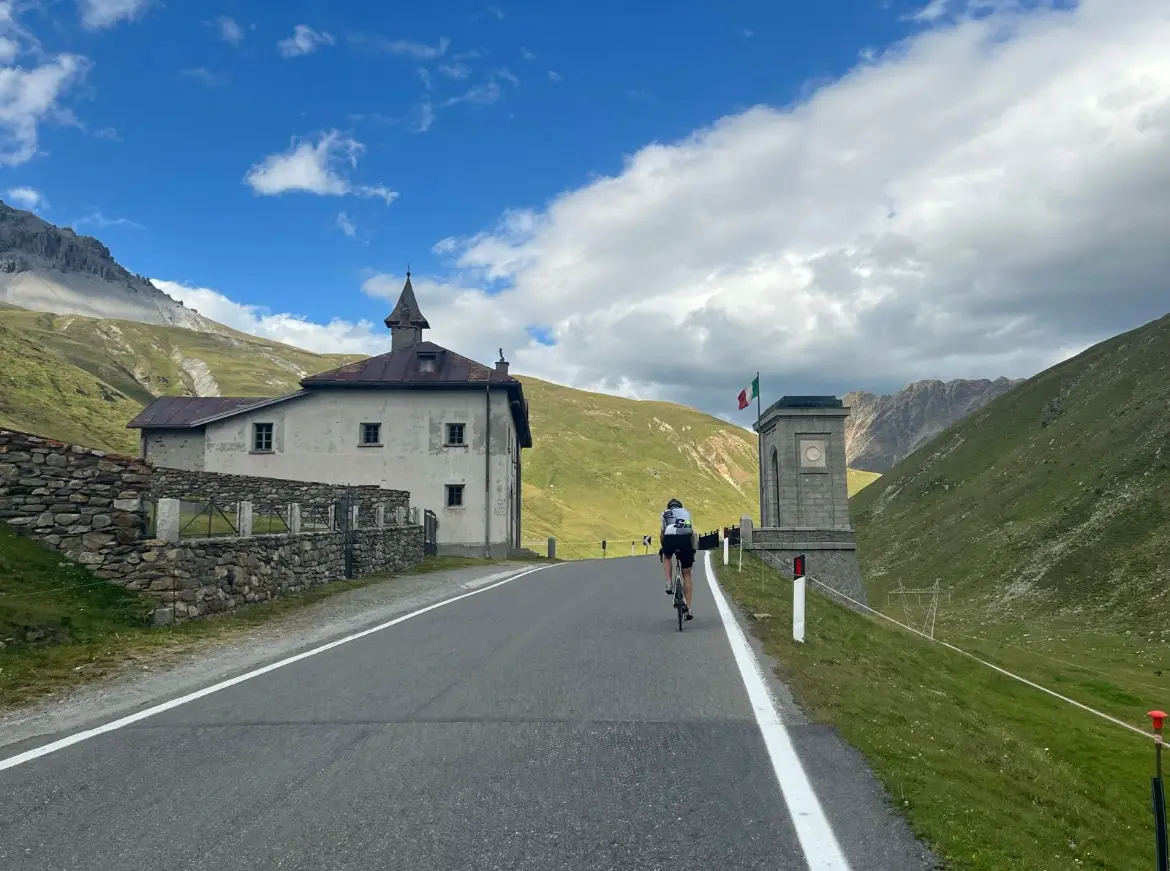 Wielrenner fietst omhoog richting de legendarische top van de Stelvio-pas langs een historisch gebouw en Italiaanse vlag.